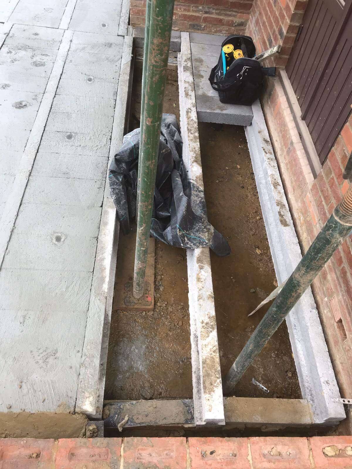 Concrete lintels and a foundation trench beside a brick wall, with a steel prop and a tool bag on site