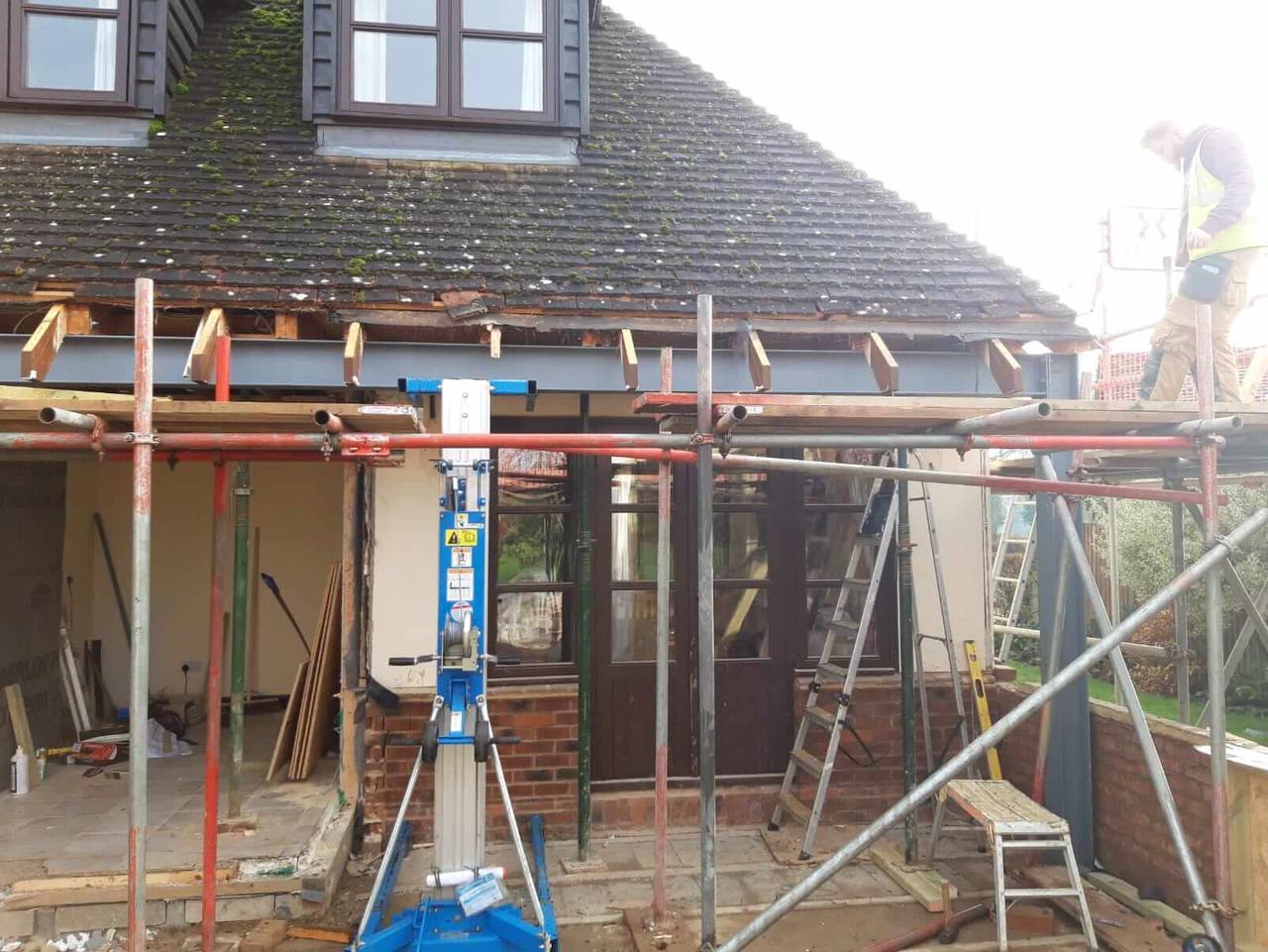 Steel beam installed across the back of a house beneath the roof line, supported by scaffolding and a material lift, ready for a rear extension