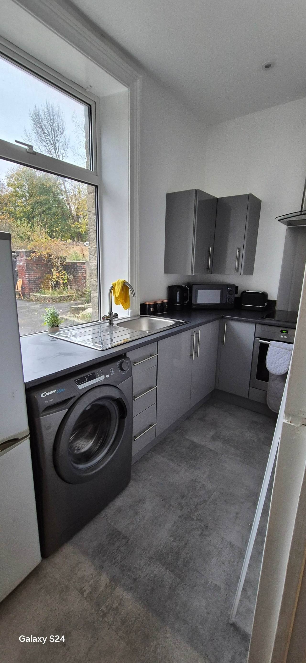 Refitted grey galley kitchen with dark worktops, a stainless-steel sink beneath a tall window and grey stone-effect flooring