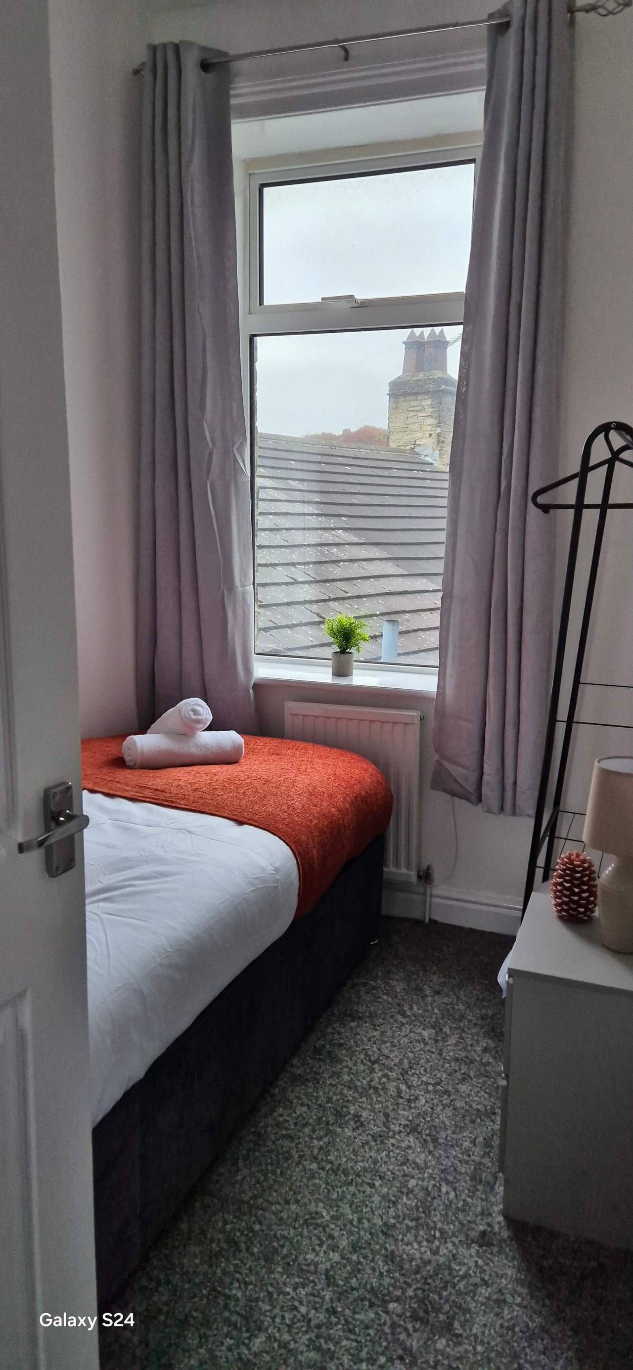 Small renovated bedroom with an orange throw on the bed and grey curtains framing a window over slate rooftops