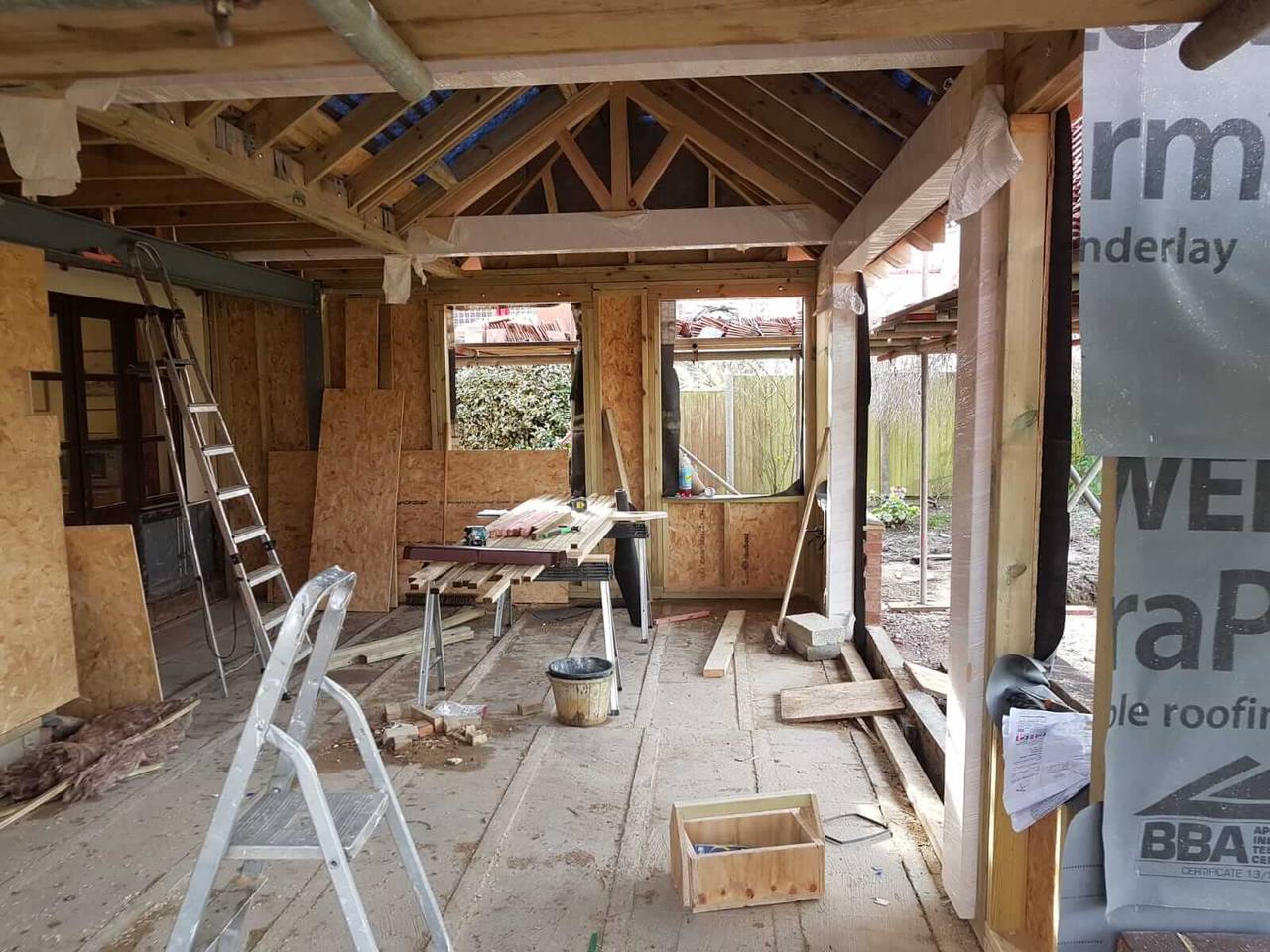 Inside a timber-framed extension under construction, with vaulted roof rafters, OSB sheathing and window openings to the garden