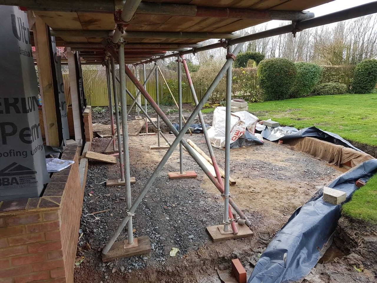 Scaffolding and props over a levelled, gravel-filled footprint beside a lawn, where a rear extension is about to be built