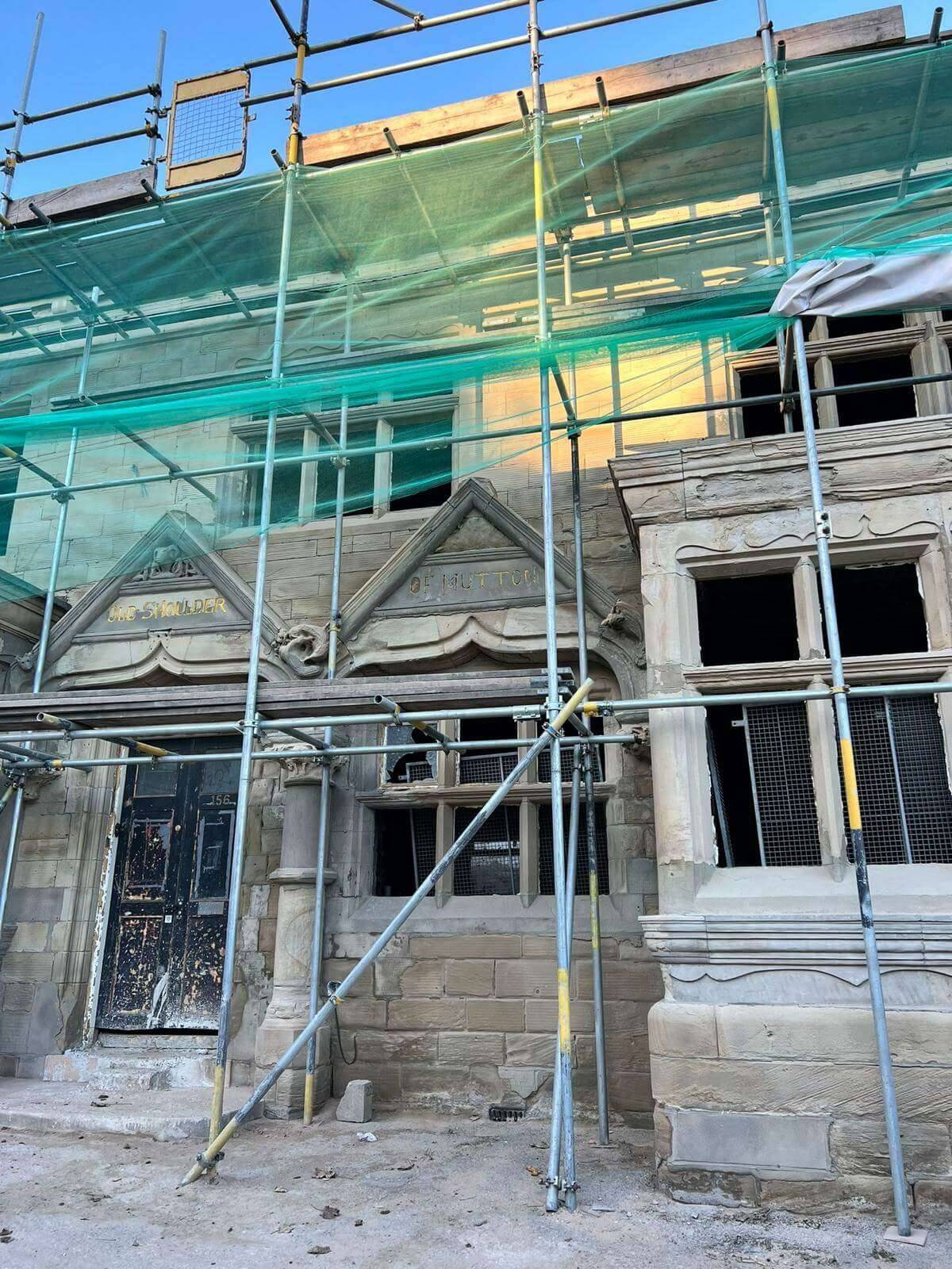 Carved stone gables and mullioned windows of a period building behind scaffolding and green netting, lit by low sun