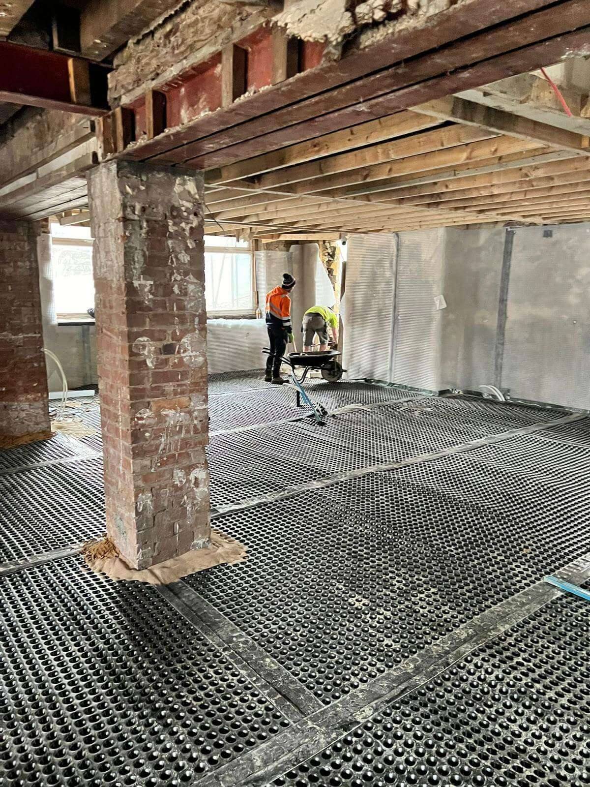 Tradespeople with a wheelbarrow working across a large ground floor covered in drainage membrane, beside a retained brick pier