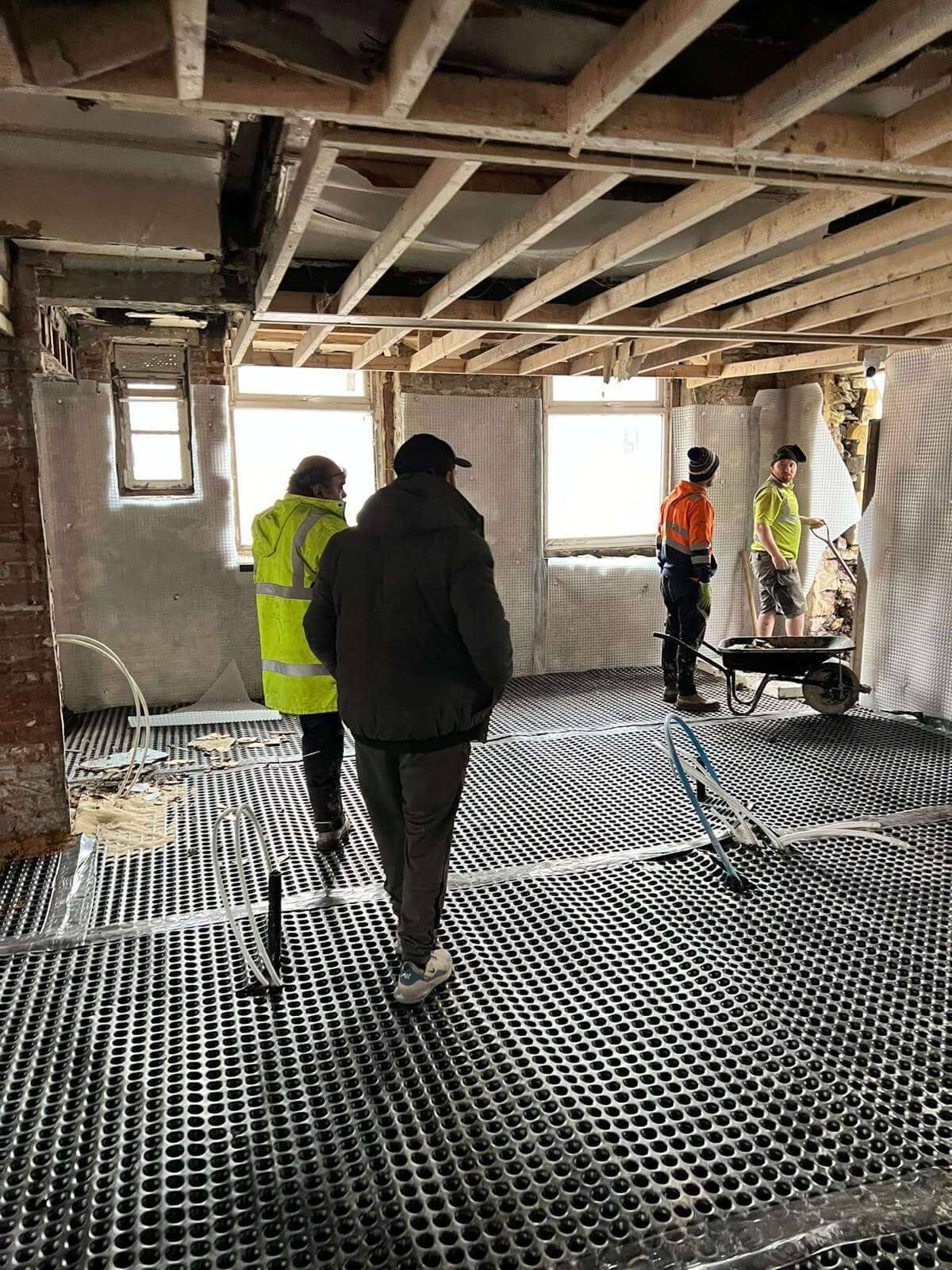 Site team walking across a stripped-back ground floor laid with cavity drainage membrane, beneath the exposed joists of the floor above
