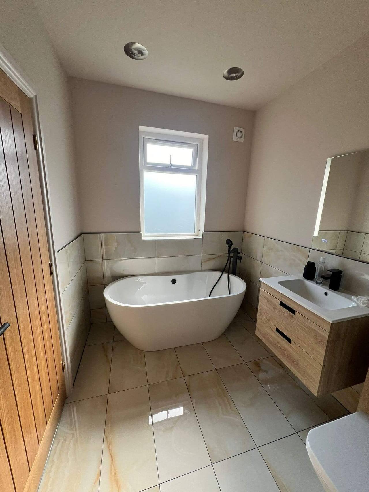 Renovated bathroom with a freestanding oval bath beneath the window, marble-effect porcelain tiles, an oak vanity unit and black fittings