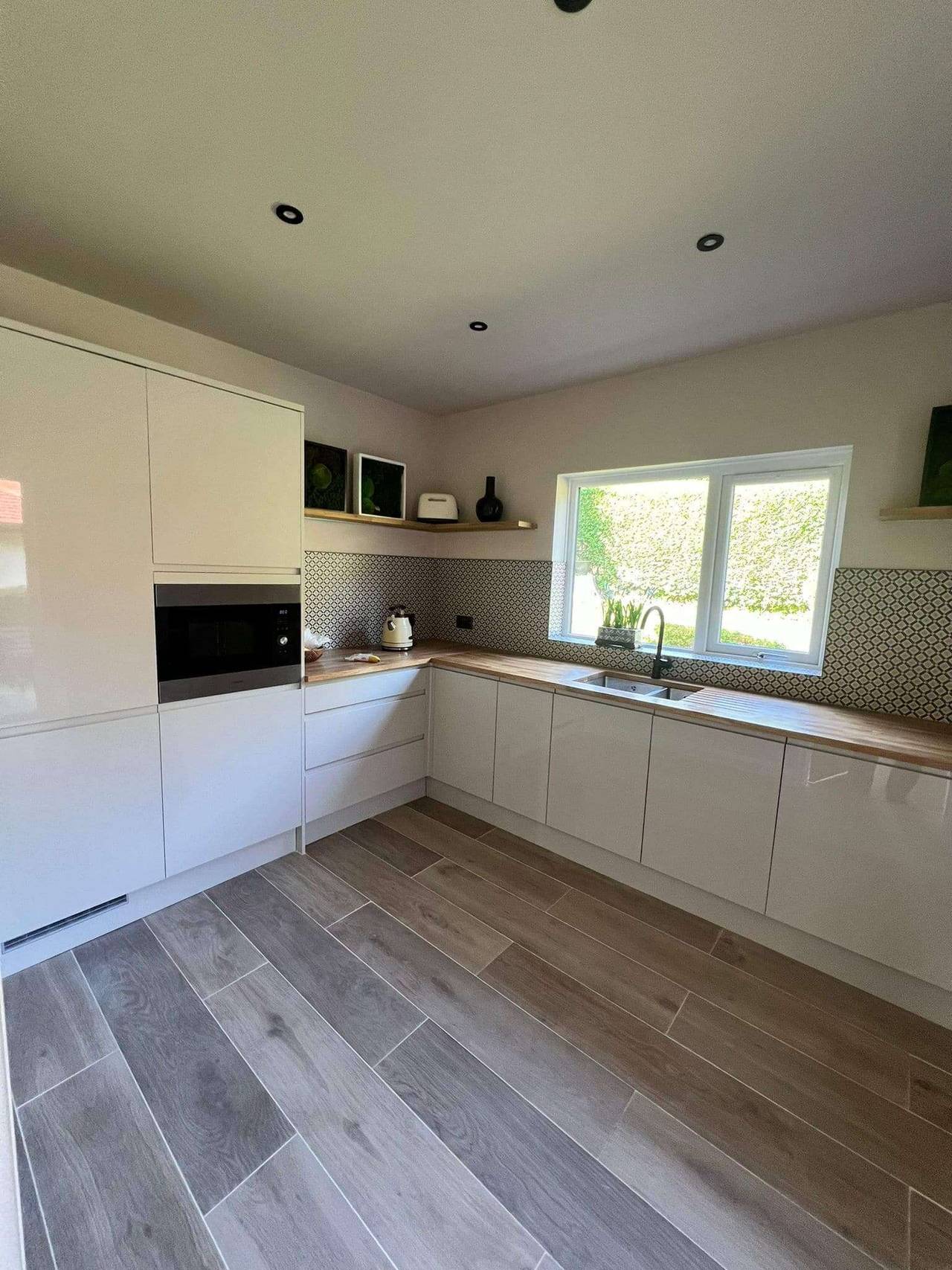 Modern handleless kitchen in white gloss with oak worktops, a patterned tile splashback, built-in oven and wood-effect tiled floor
