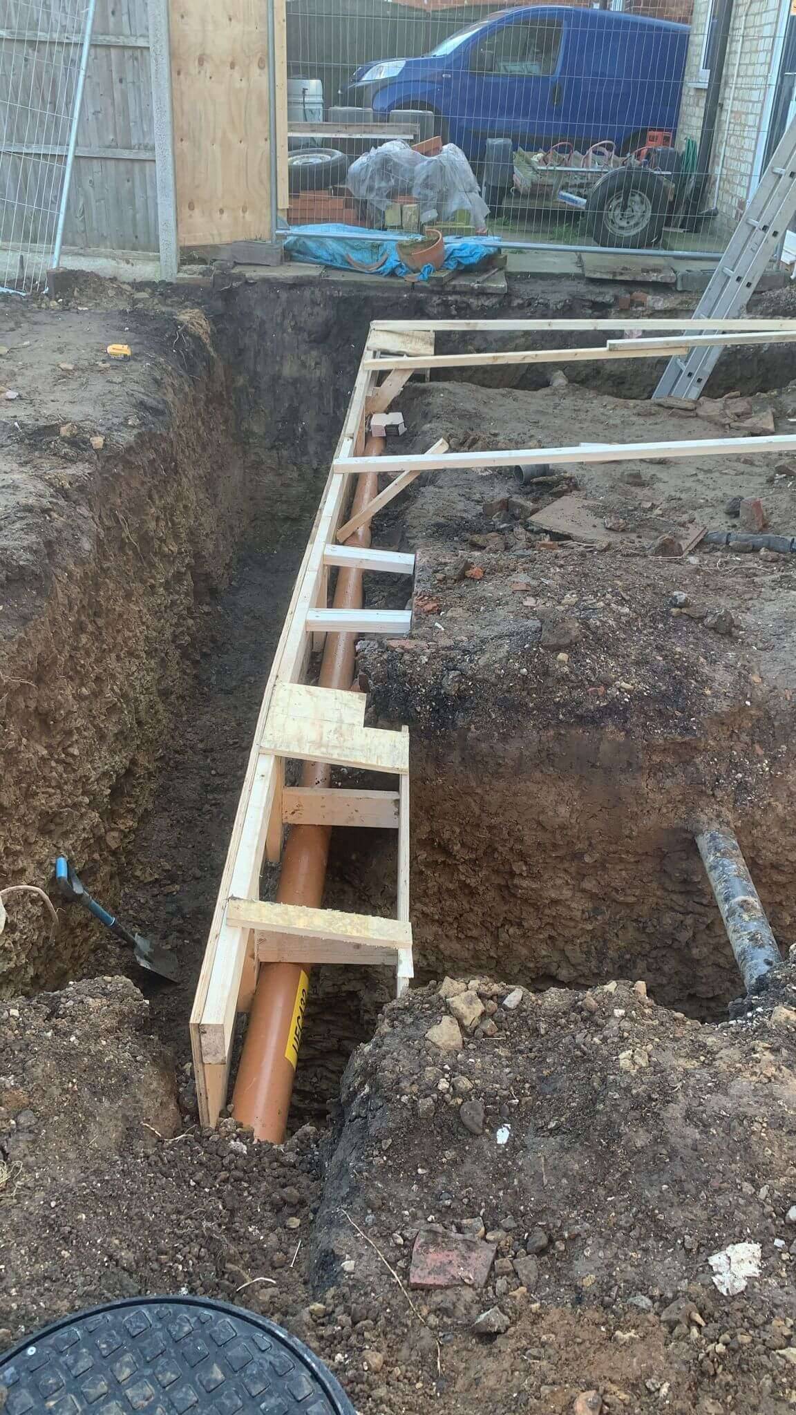 Extension foundation trench with timber shuttering and a new orange drainage pipe running alongside