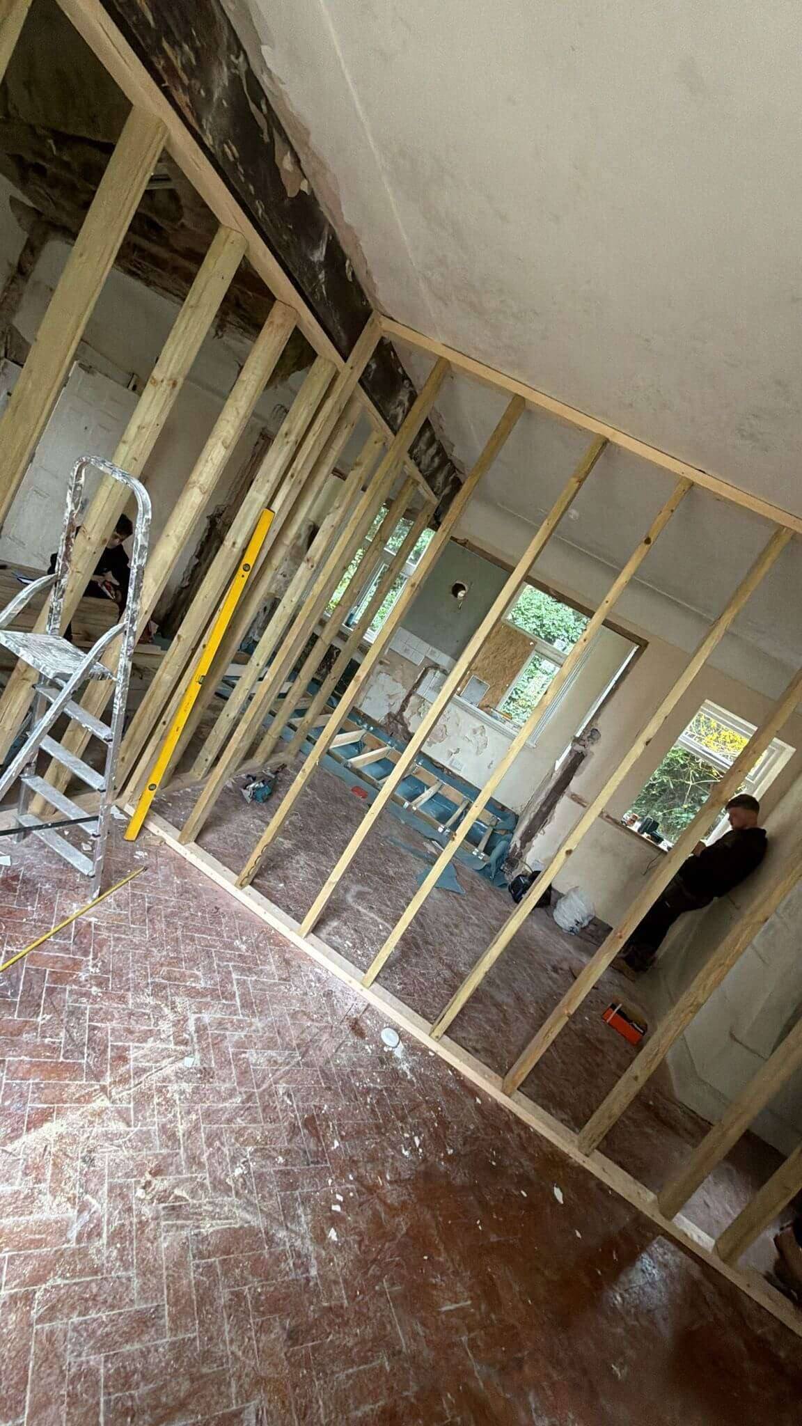 Timber stud partition going up across a large room with a herringbone brick floor, with tradespeople working in the background
