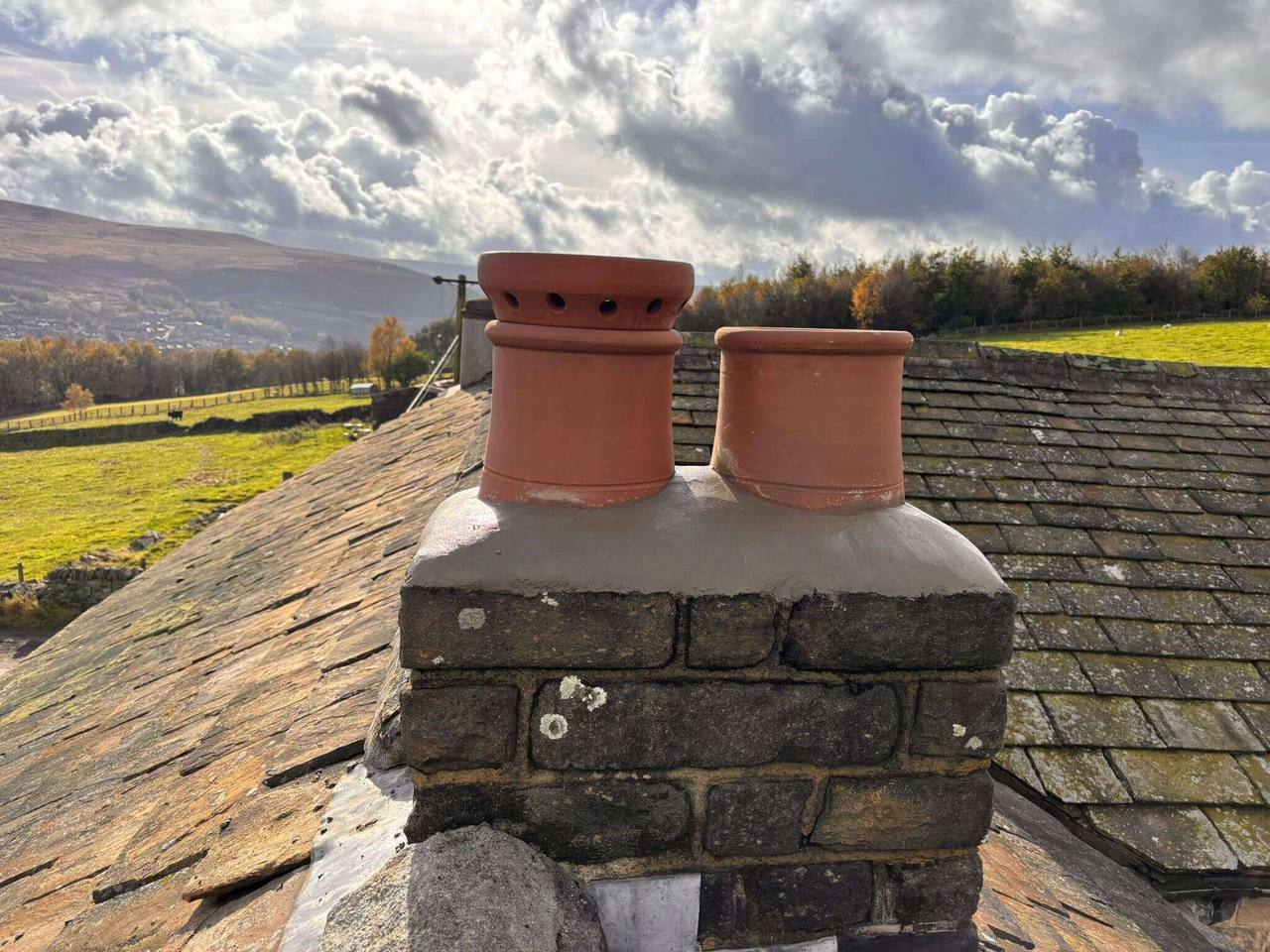 Rebuilt stone chimney stack with new terracotta pots on a stone slate roof, overlooking Pennine hills under a dramatic sky