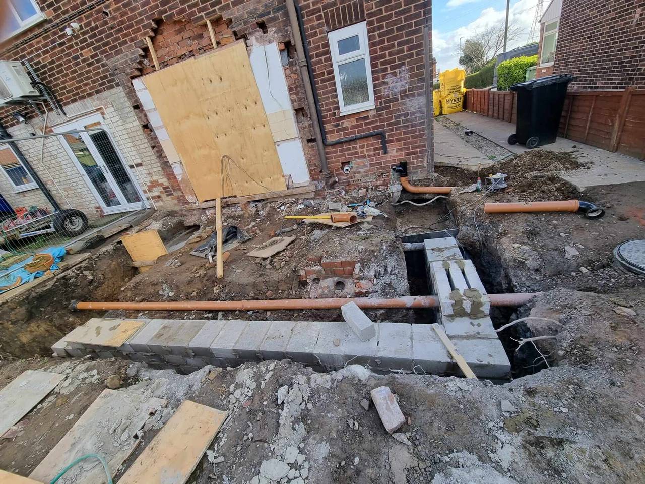 Groundworks for a side extension, with blockwork footings, new orange drainage pipes and a boarded-up opening in the brick house wall