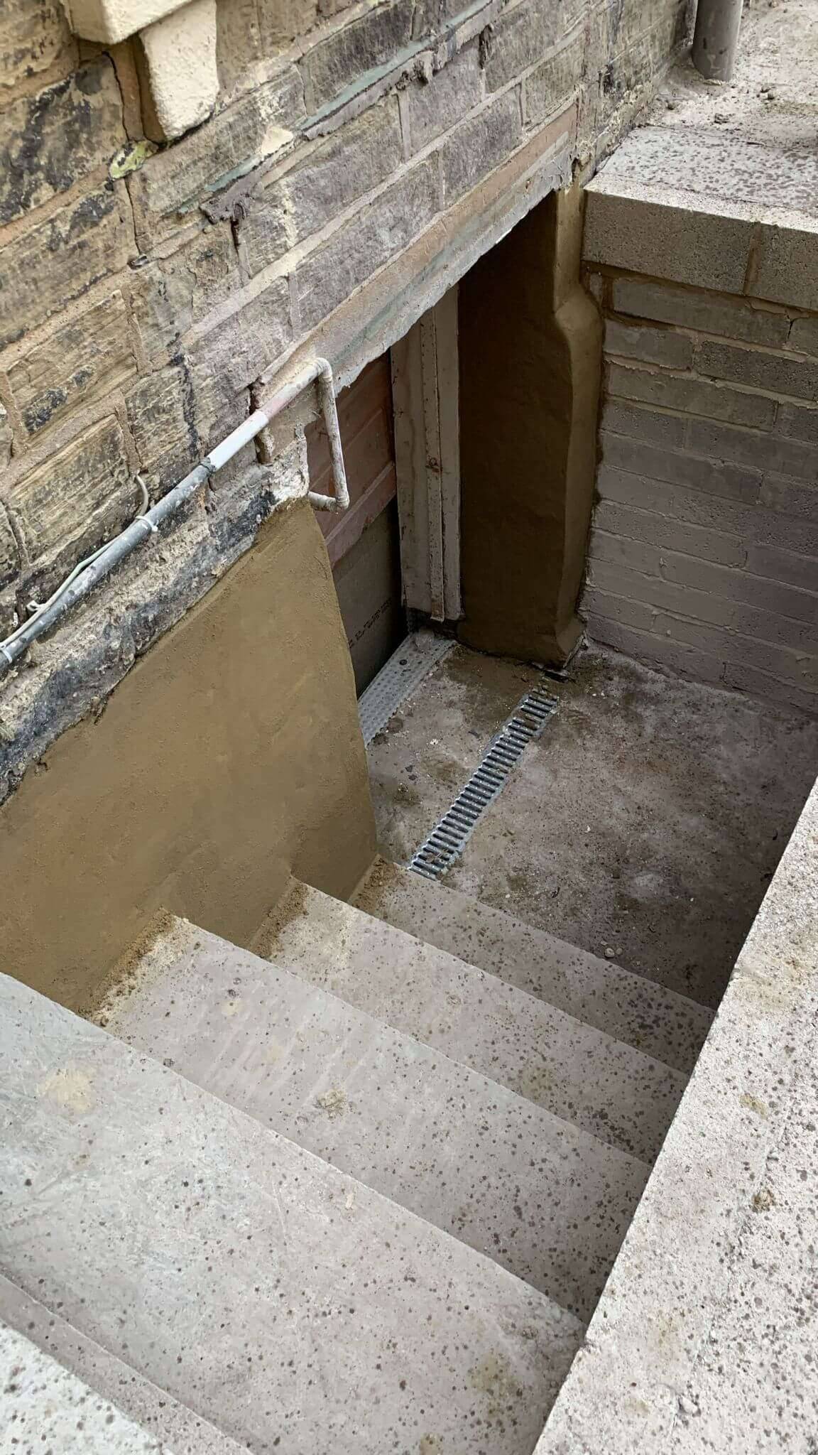 New concrete steps leading down to a rendered basement entrance with a drainage channel, beside a stone wall