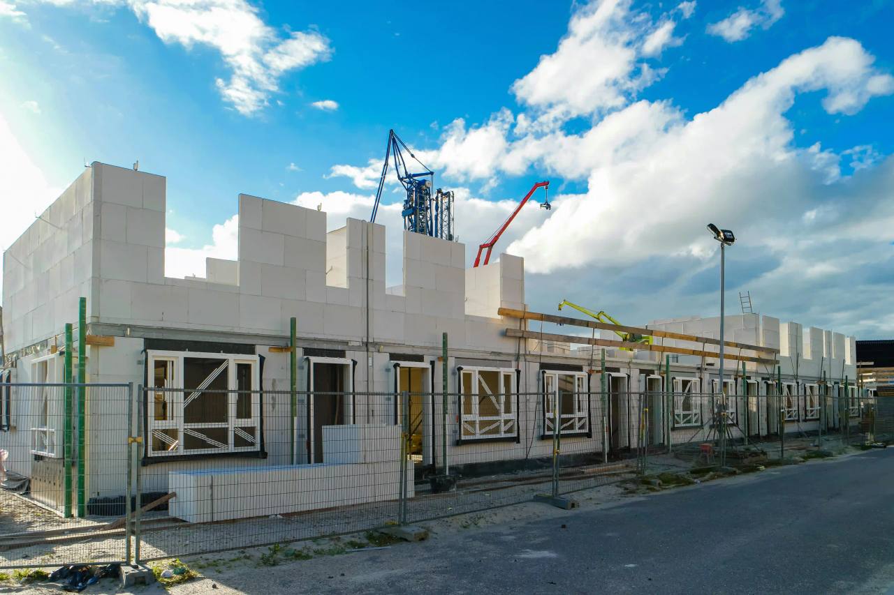 Terrace of new homes under construction in white blockwork, with window frames fitted and cranes beyond, under a blue sky